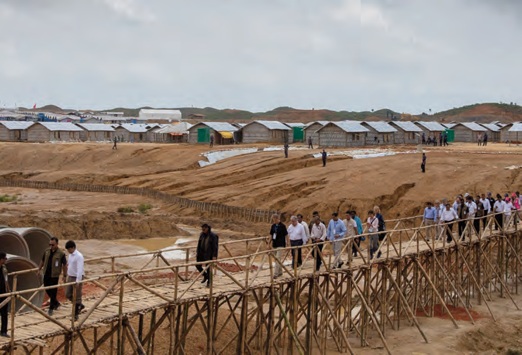 A delegation crosses a bamboo walkway through a sprawling refugee camp
