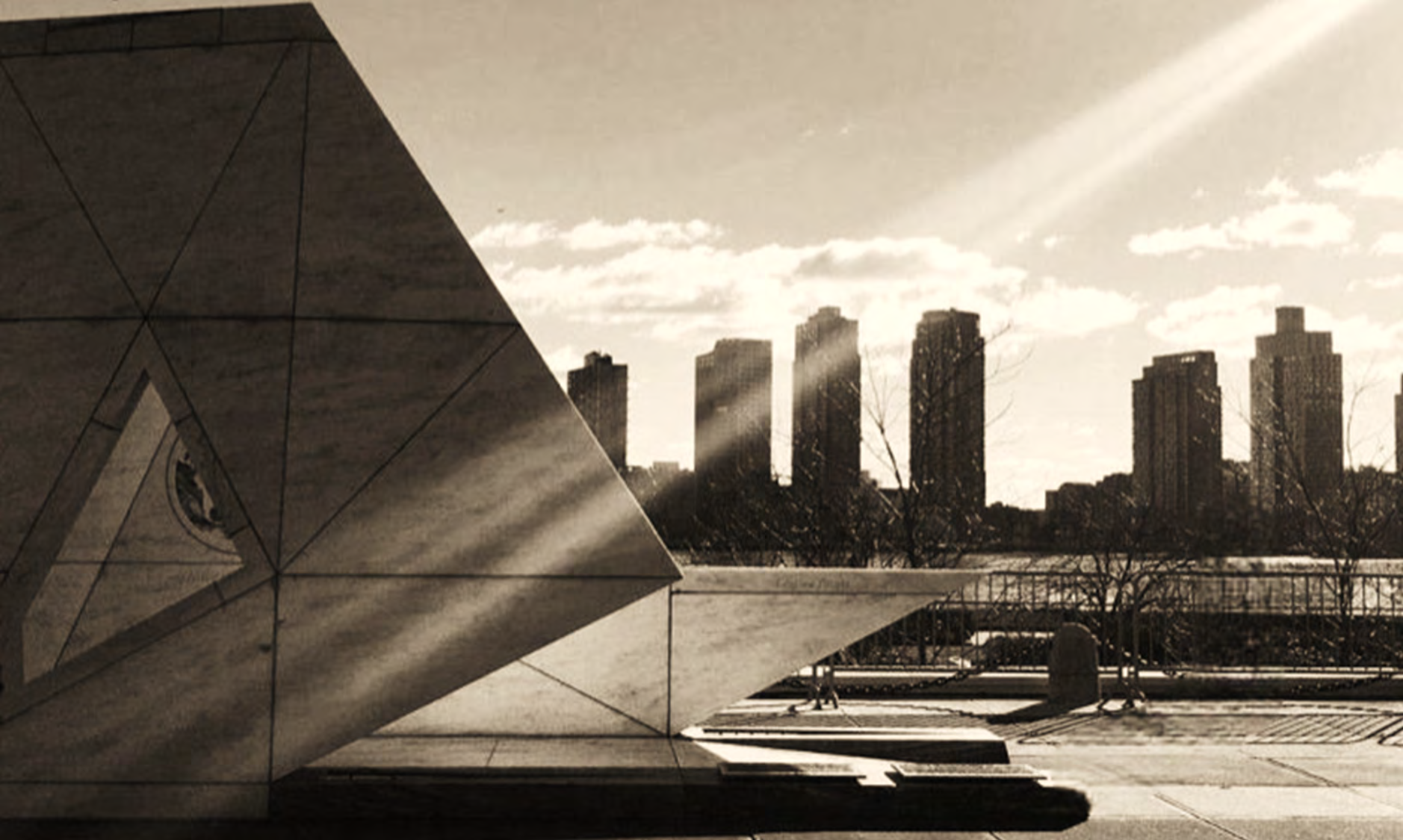 A sepia photograph of an angular marble monument with a city skyline behind it