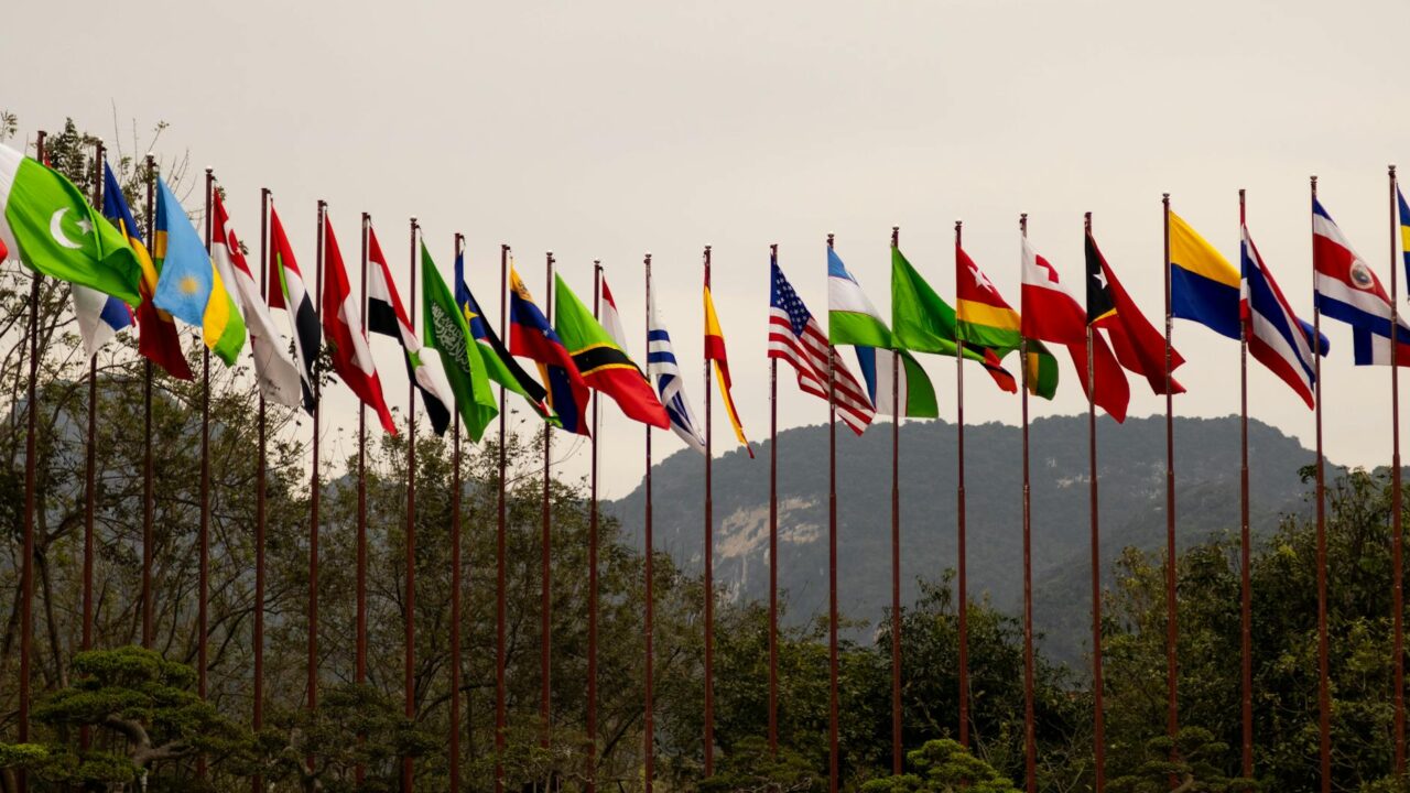 A row of flags from various countries on poles in front of a mountainous backdrop.