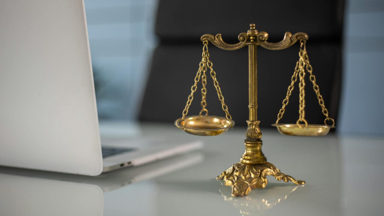 A golden scale of justice on a desk with a laptop and papers in the background.