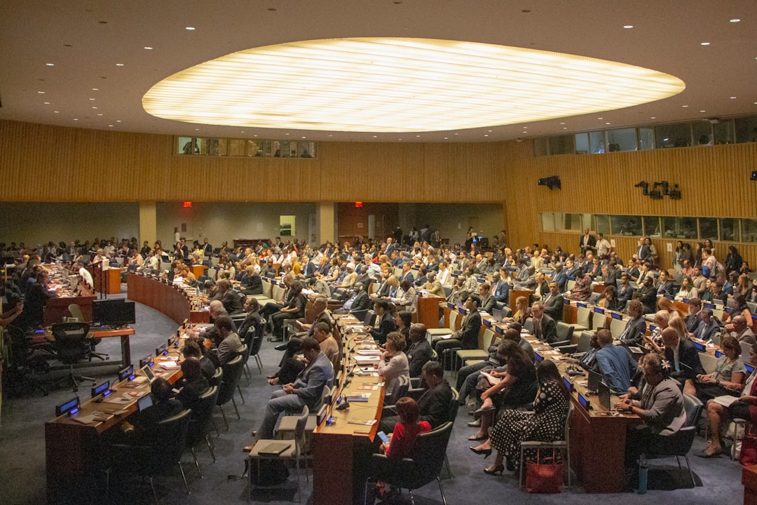 The image depicts a crowded conference room at the United Nations headquarters, with delegates seated around a circular
