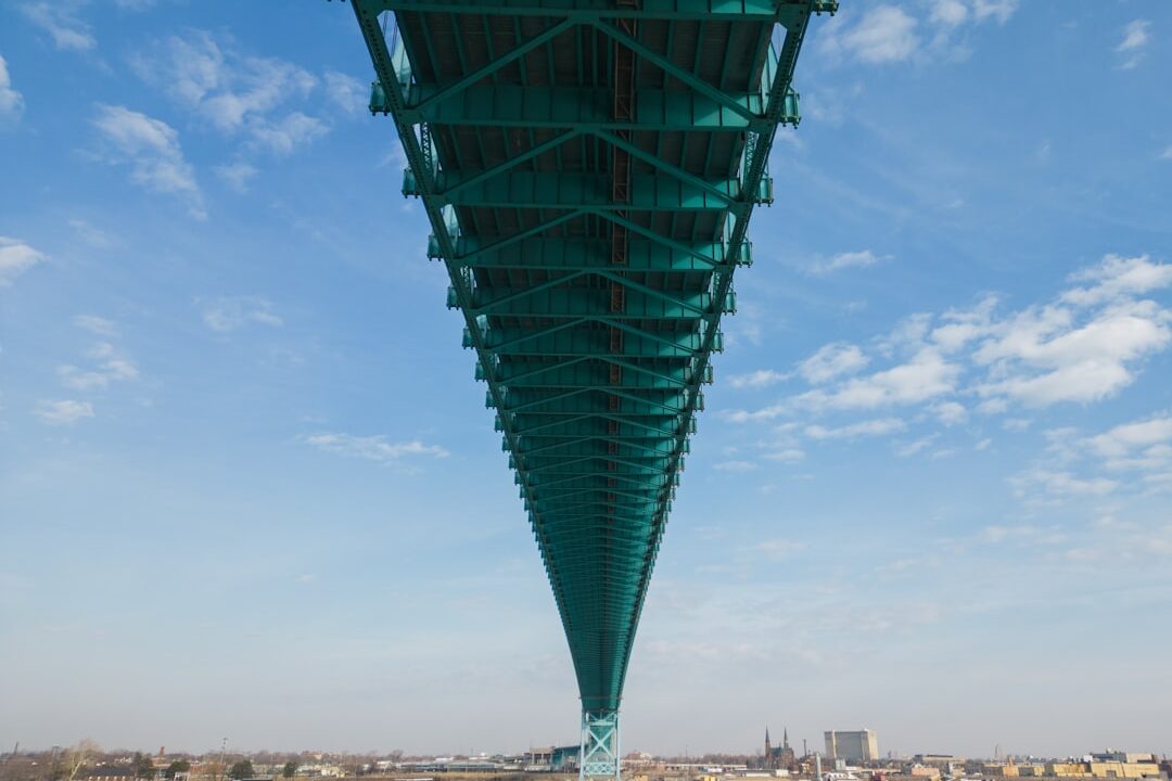 The image depicts a large green suspension bridge spanning over a body of water, with the city skyline in the background