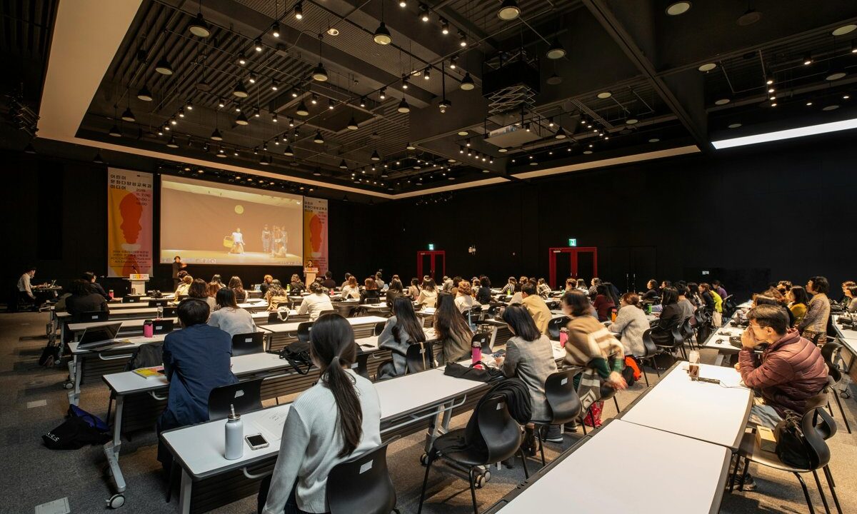 A conference hall with rows of attendees seated at tables facing a stage and screen, a clean meeting scene standing in for the in-person CGS Annual Meeting.