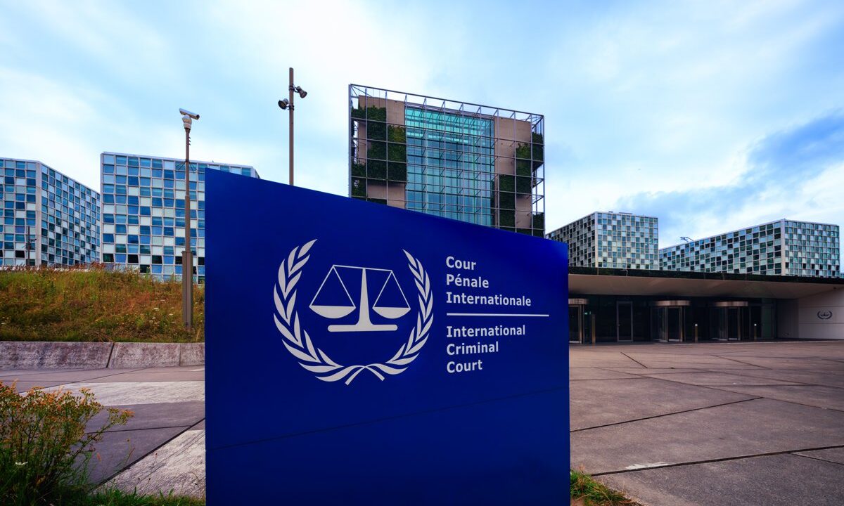 The International Criminal Court complex in The Hague, with the court's sign (scales of justice and the words Cour Penale Internationale / International Criminal Court) in the foreground and the full building behind, a definitive establishing shot of the institution.