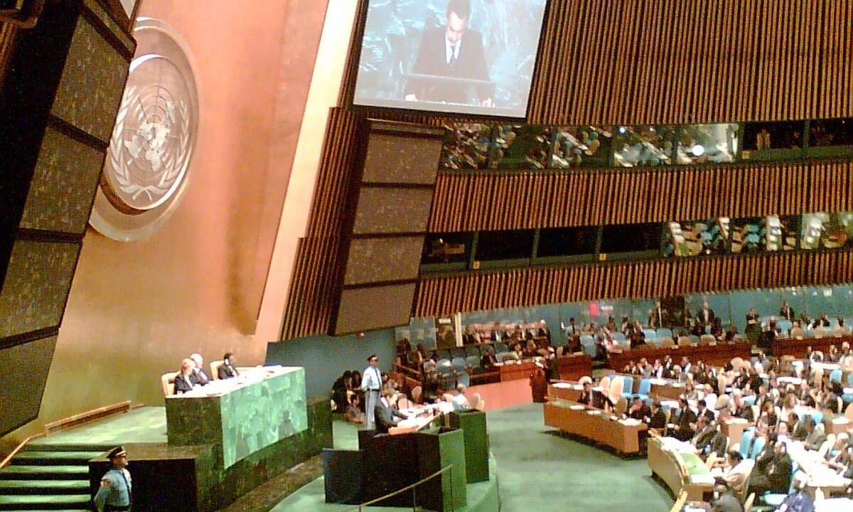 The United Nations General Assembly in session, with a speaker at the green marble rostrum beneath the UN emblem and delegates filling the hall: the body that adopted the UN75 modalities resolution.