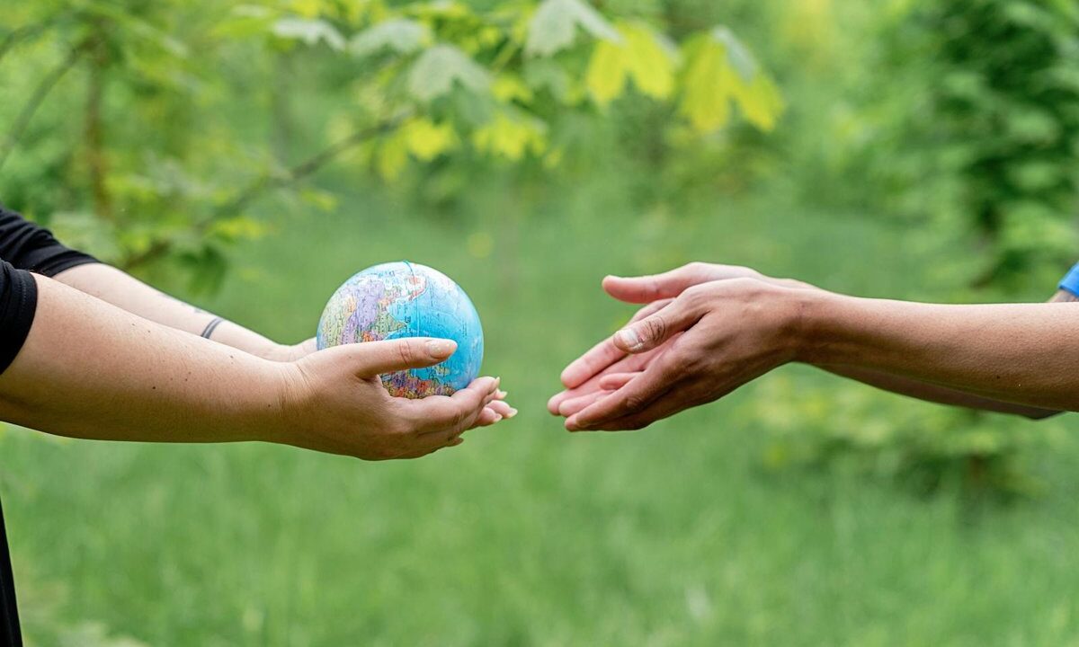 Two hands passing a small globe to another pair of hands against a green background, a conceptual image of legacy, stewardship, and handing the world on to the next generation.