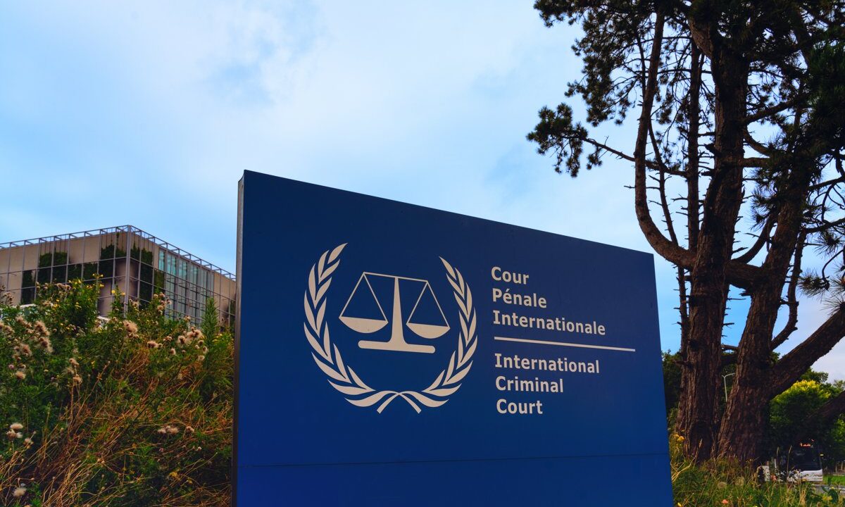 A close view of the International Criminal Court sign in The Hague, the scales-of-justice emblem and the court's name in French and English dominant, with the building behind.