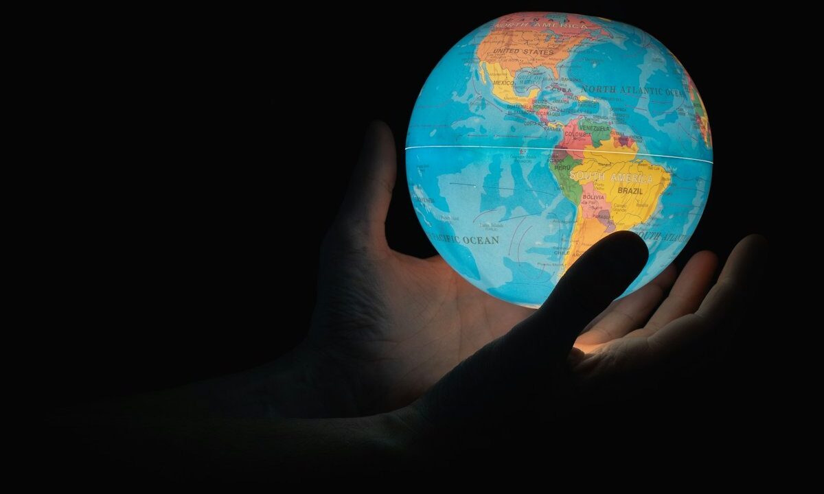 A colorful illuminated world globe (the Americas facing forward) cradled in an open human hand against a black background: a conceptual image of holding the whole world and taking responsibility for it, matching an essay on solving global problems together.