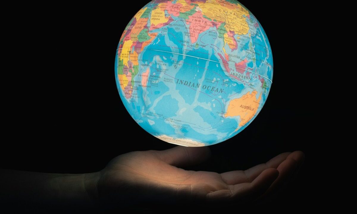 A glowing political globe of the whole Earth resting above an open hand against black, a clean conceptual image of one unified world.