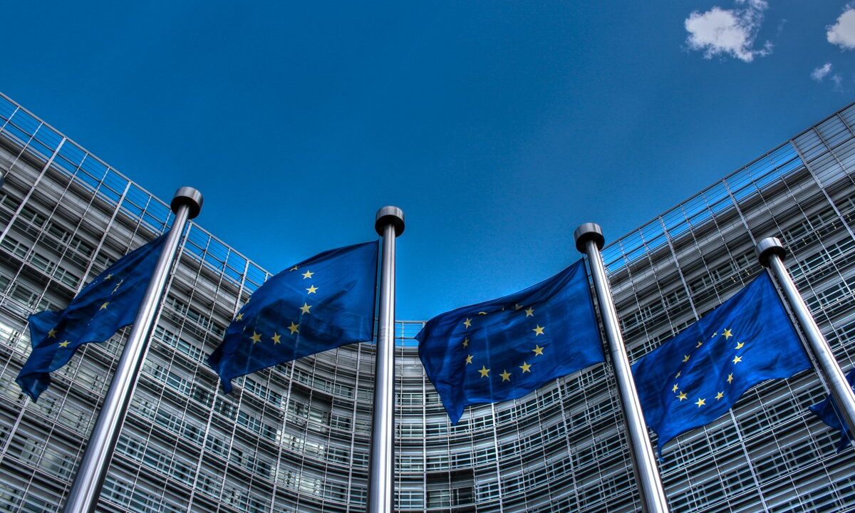 A row of European Union flags before the curved facade of the Berlaymont (European Commission), a clean emblem of supranational European federation set against national sovereignty.