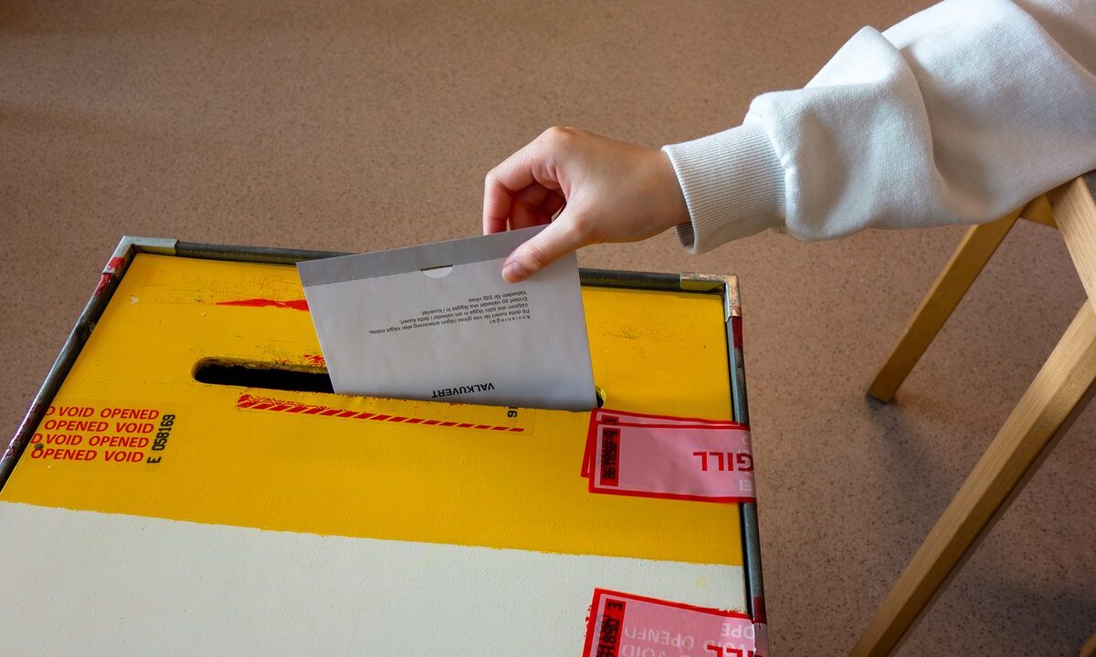 A hand dropping a paper ballot into a sealed, tamper-evident ballot box at a polling station: the concrete act of democratic voting.
