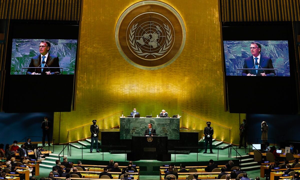 The United Nations General Assembly in full session, head on toward the gold emblem with a speaker at the rostrum and delegates filling the floor, the body whose member contributions fund the UN.