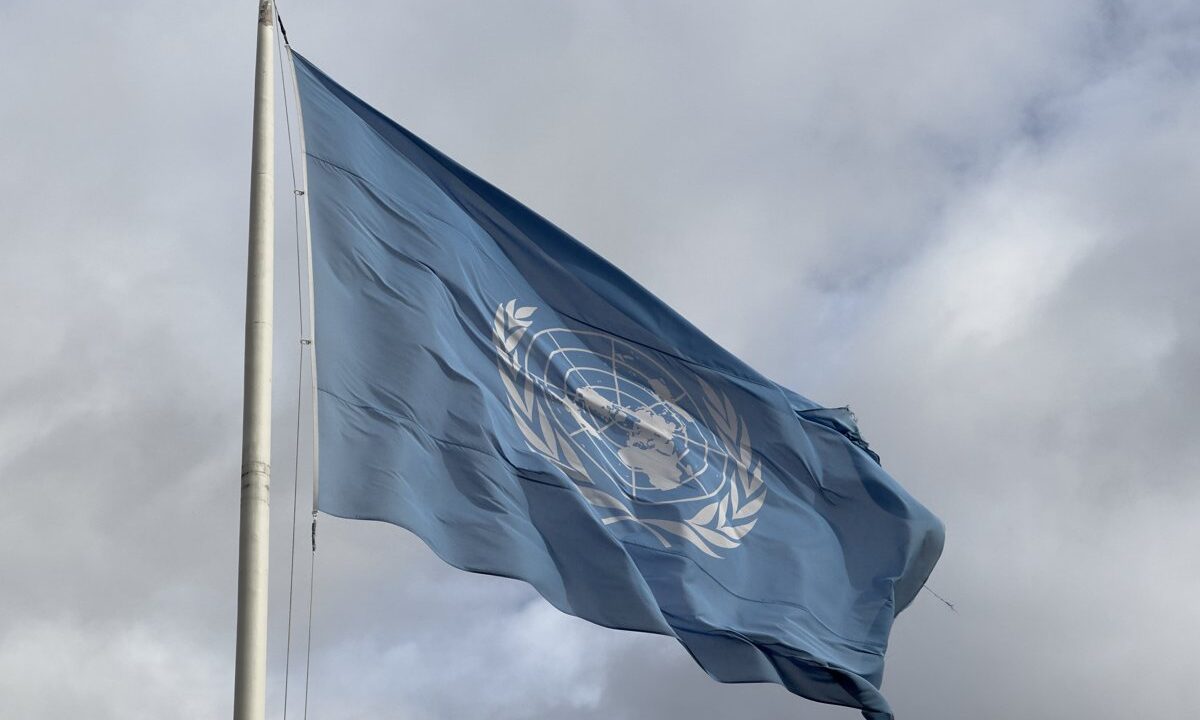 The flag of the United Nations, light blue with the world map and olive branches emblem, waving against a cloudy sky.