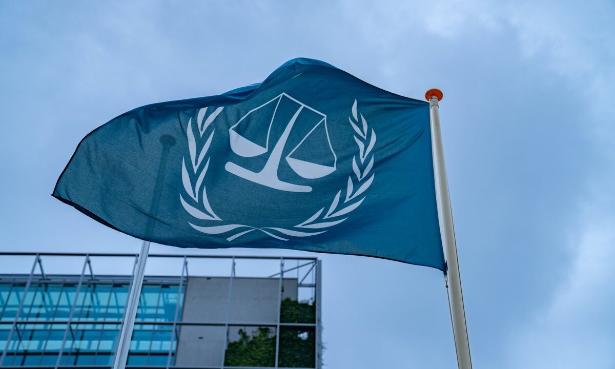 The flag of the International Criminal Court (scales of justice wreathed in laurel) flying outside the court in The Hague, representing the Rome Statute regime the article argues the United States should join.