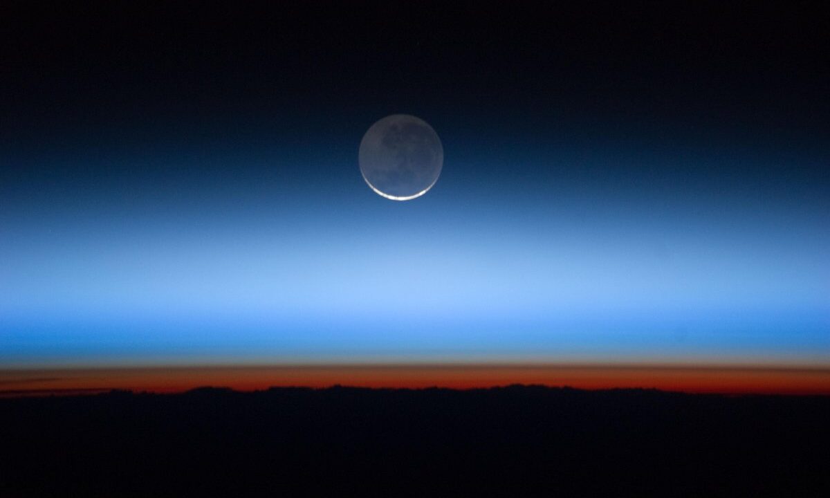 Earth's thin atmosphere seen edge-on from the International Space Station, the fragile blue shell fading to the orange of sunset with the Moon above: a direct visual metaphor for planetary boundaries and a safe operating space for humanity.