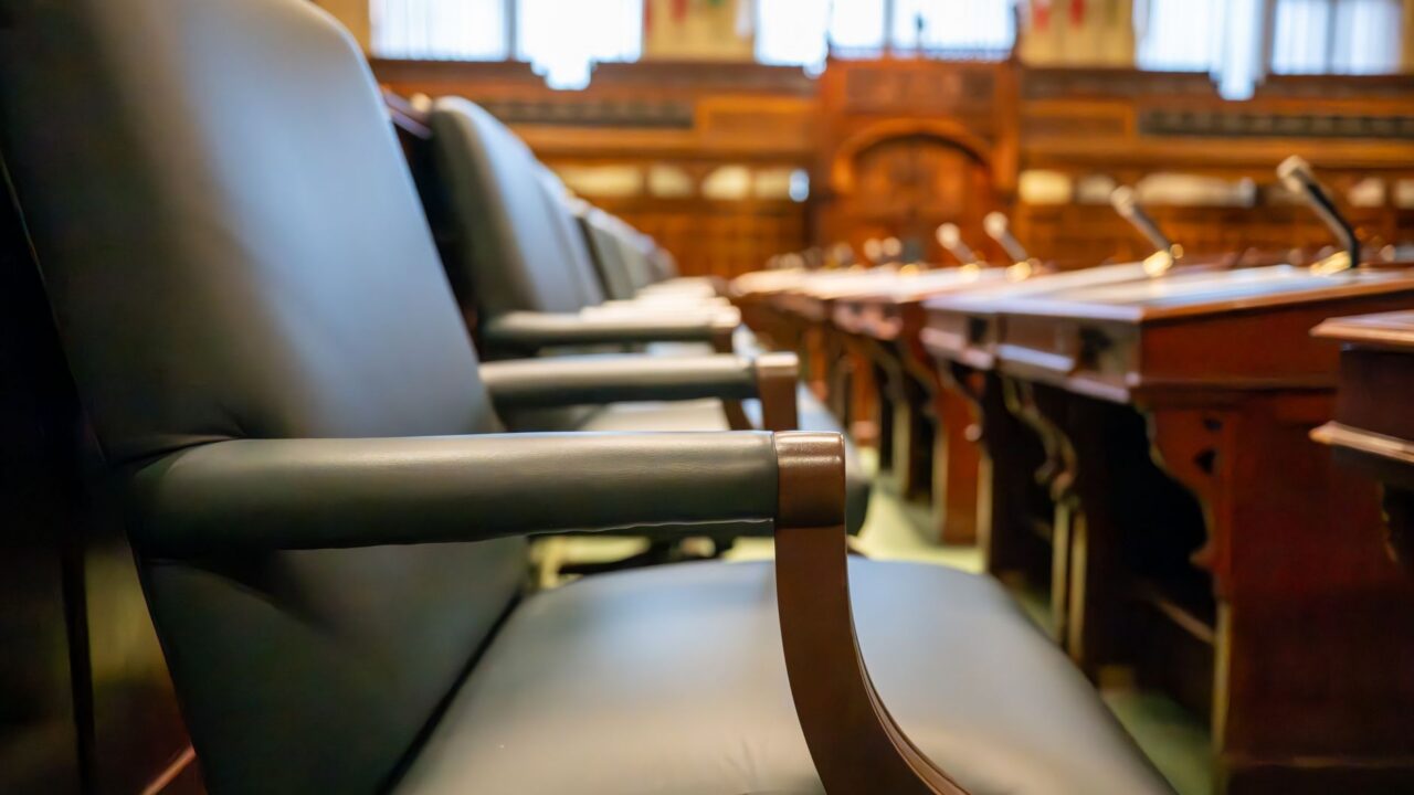 The image shows the interior of a legislative chamber, with rows of leather-upholstered desks and chairs facing a centra
