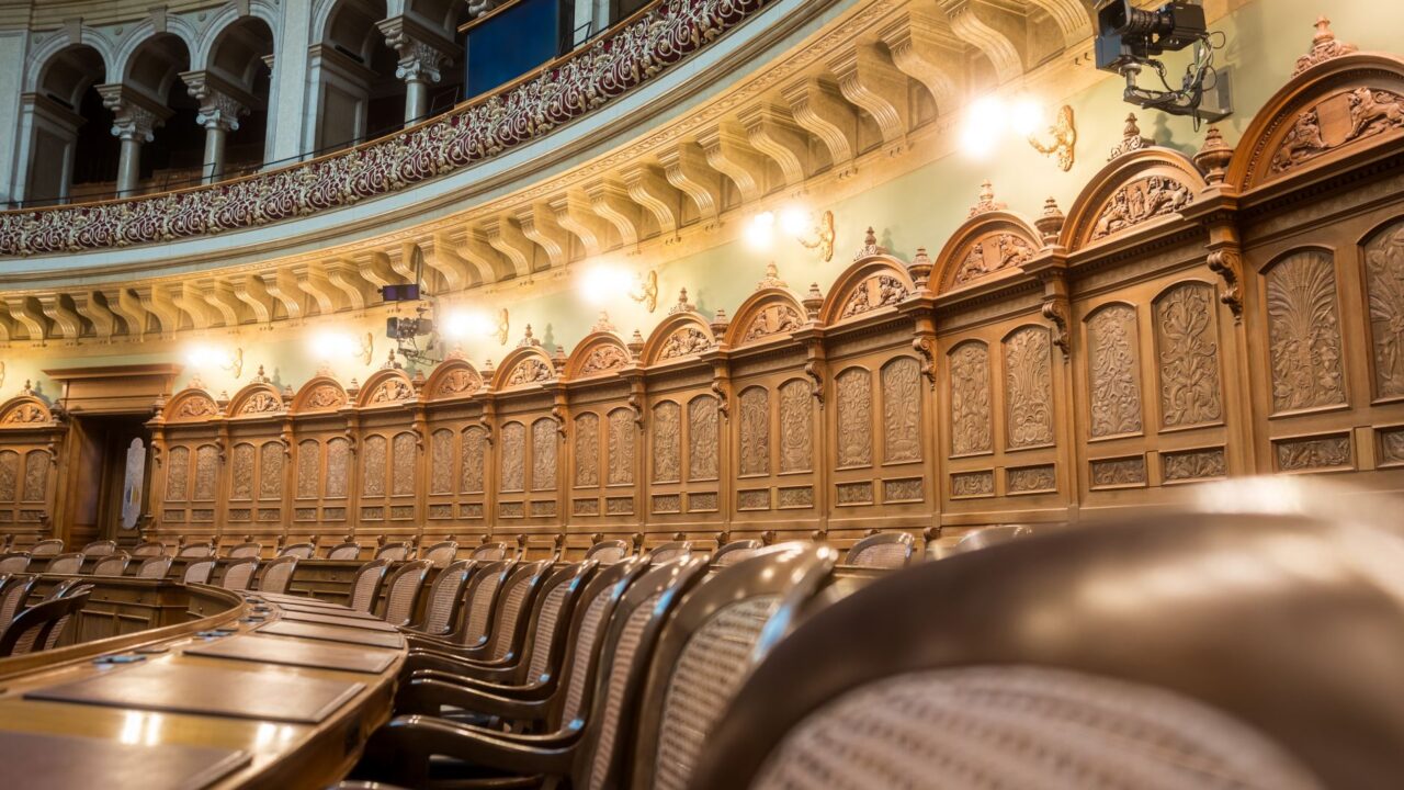 The interior of a formal legislative chamber with rows of wooden benches and a high ceiling with ornate decorations.