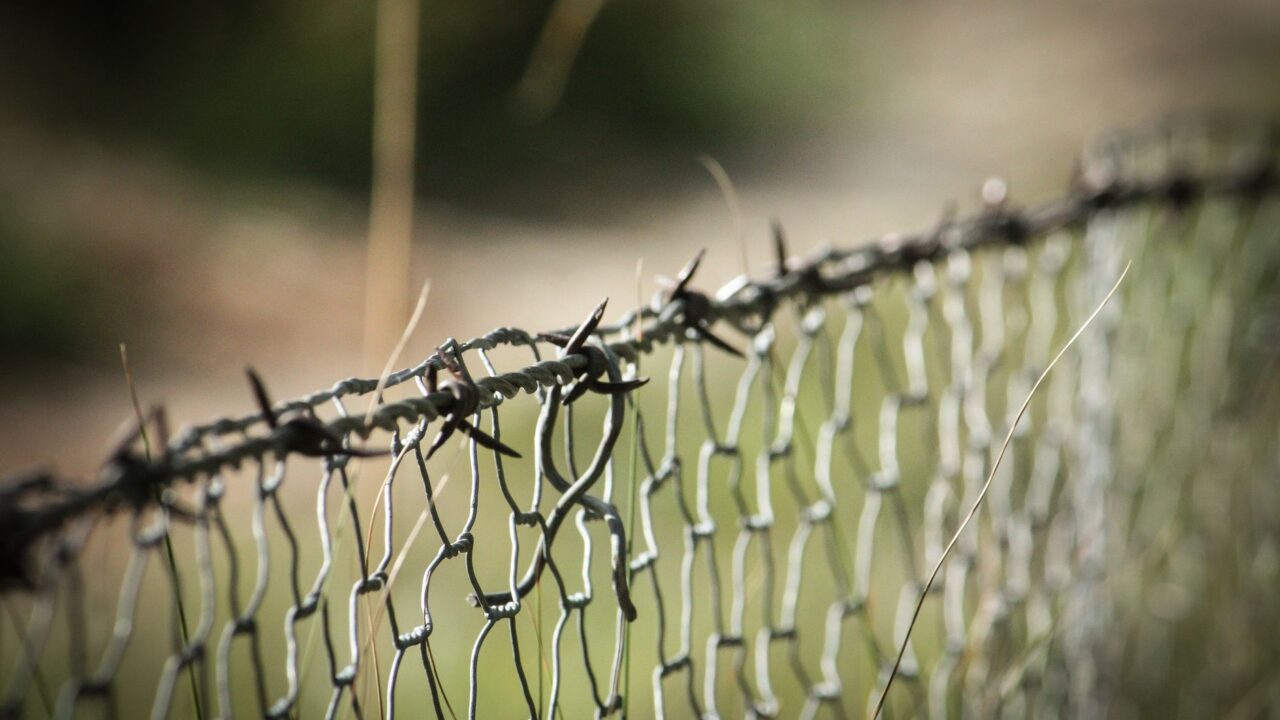 Barbed wire along a chain link fence in front of a grassy field.