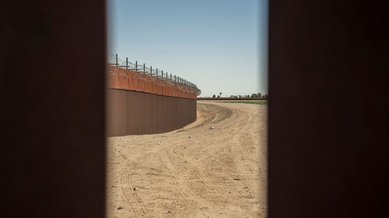 A view through a fence showing a construction site with a barrier wall under a clear sky.