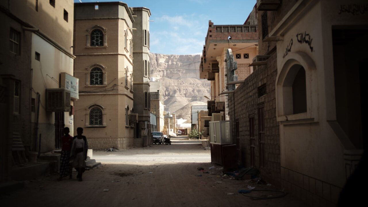 A narrow street of tall stone buildings beneath rocky cliffs.