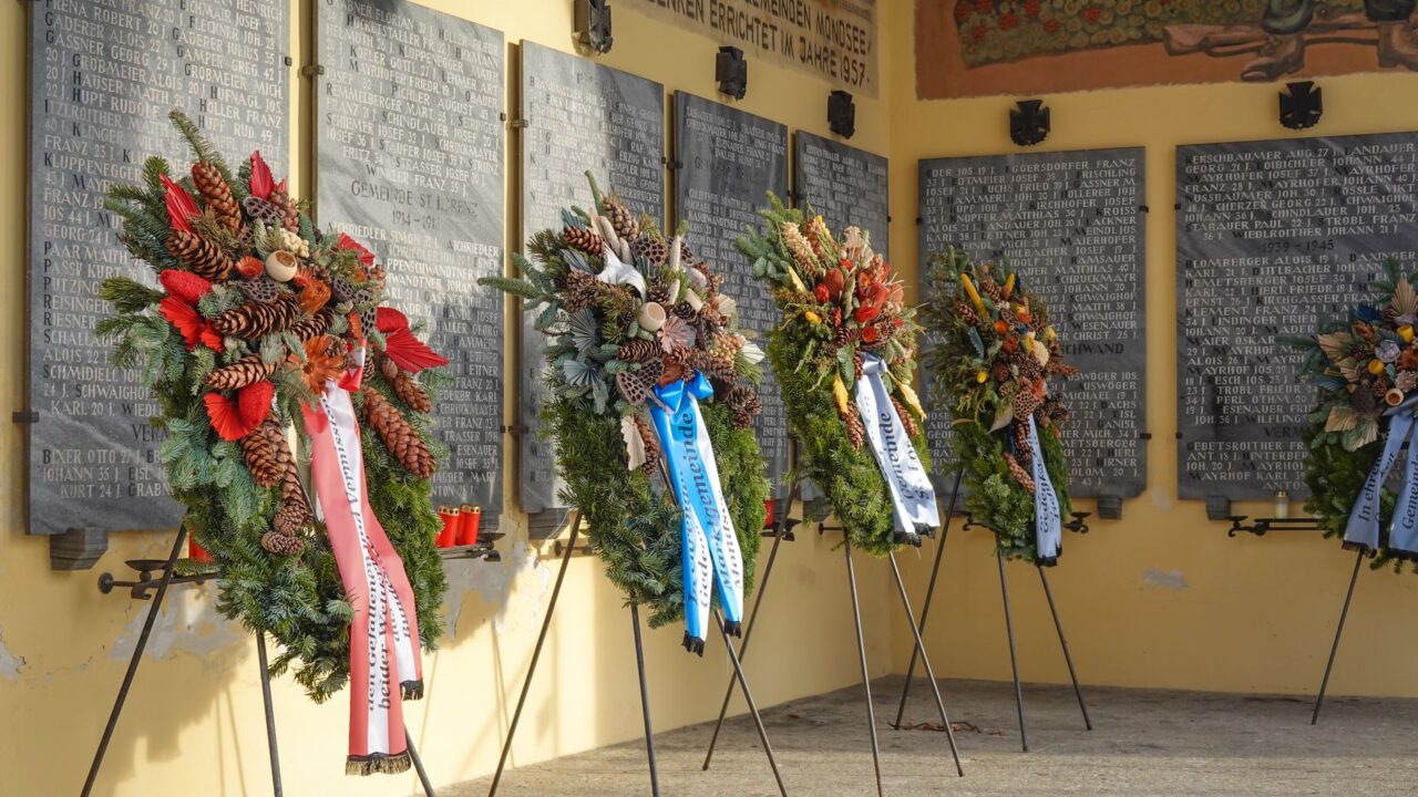 Flower wreaths on stands before stone memorial plaques.
