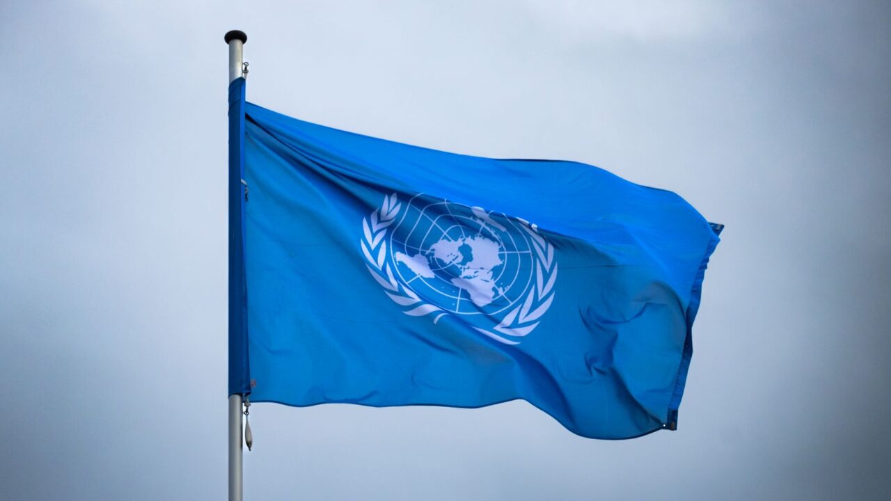 The blue United Nations flag flying from a pole under a grey sky.