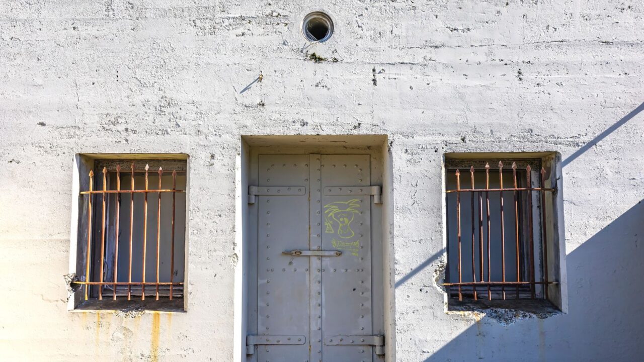The image shows a door to a prison cell with barred windows and a concrete exterior.
