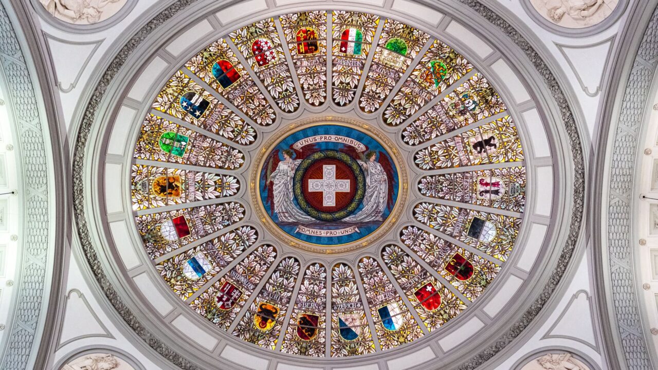 The image shows the interior of a church with a large, ornate stained glass ceiling featuring a cross at its center, sur