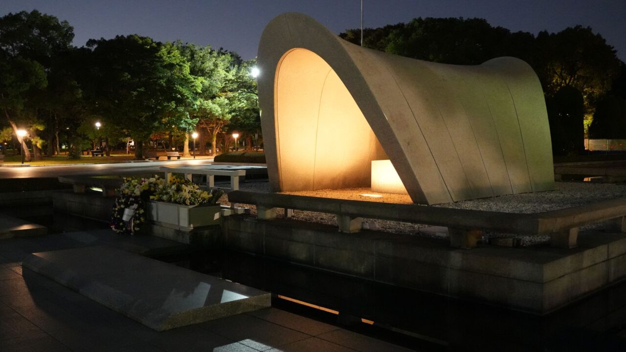 The photograph shows a modern architectural memorial at night, illuminated by artificial lights, with a reflective surfa