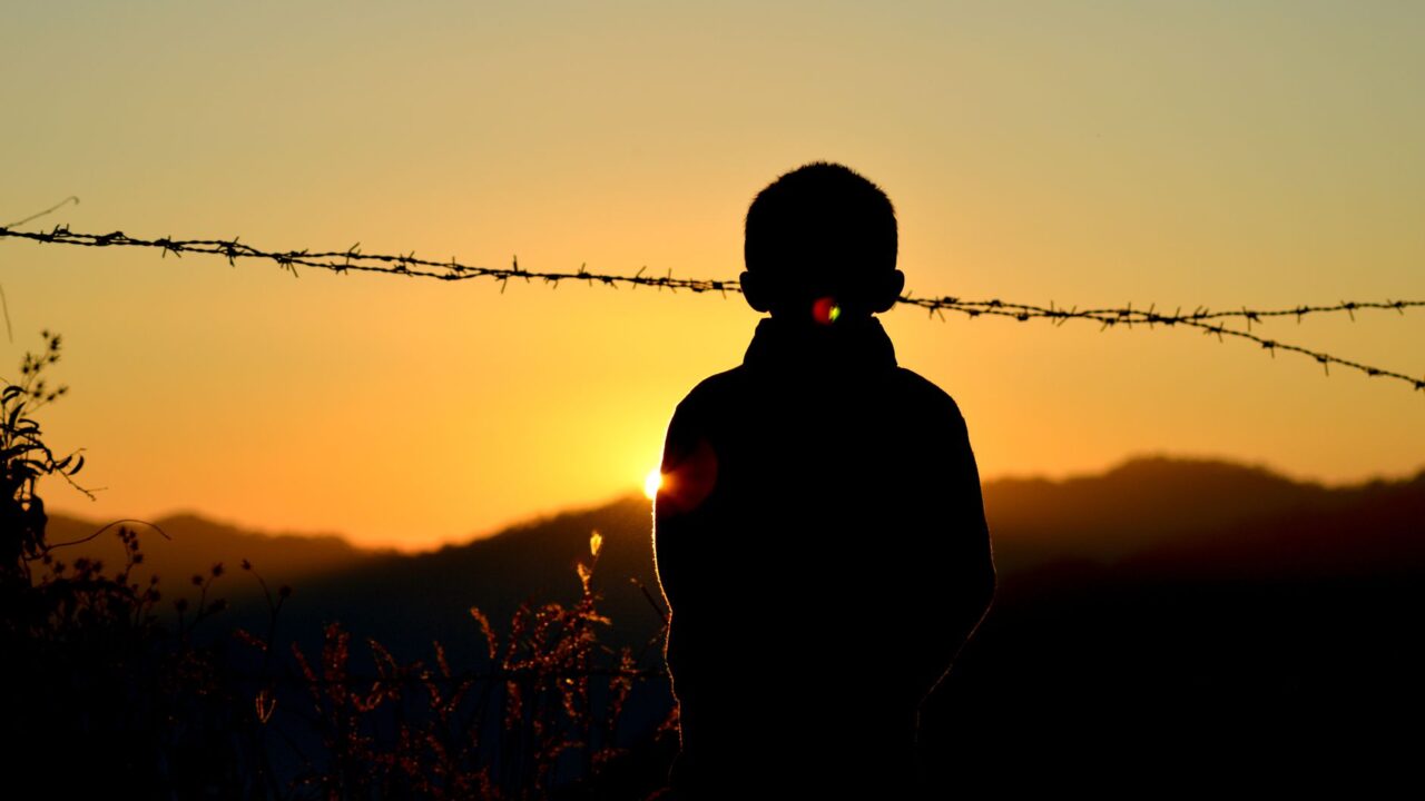 The silhouette of a child behind barbed wire at sunset.