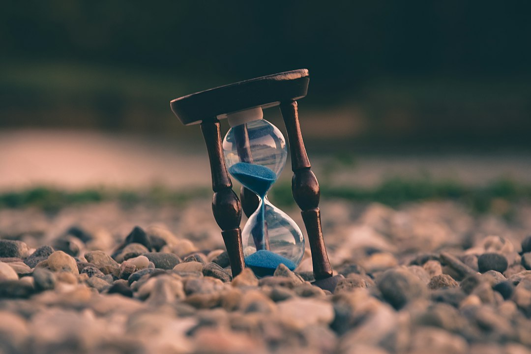 An hourglass sits on a gravel surface, with the sand running out, symbolizing the passage of time.