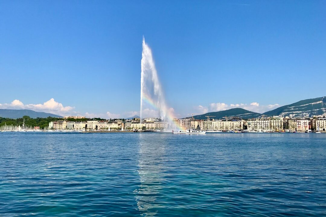 A fountain spraying water in the middle of a city, with a clear blue sky and mountains in the background.