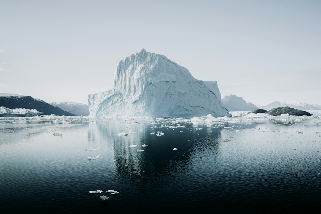 A large iceberg floats in the ocean, with a mountainous landscape in the background.
